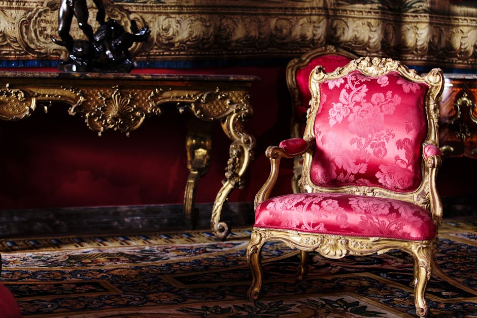 A richly upholstered antique armchair featuring ornate carved wooden legs and a patterned fabric with red, gold, and beige tones, positioned in a luxurious room near a large window with a rounded top.