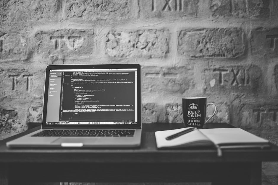 A black-and-white photo of a workspace featuring a laptop displaying a coding interface on a wooden desk, with a mug bearing the phrase 'Keep Calm and Drink Coffee' placed next to a closed notebook an