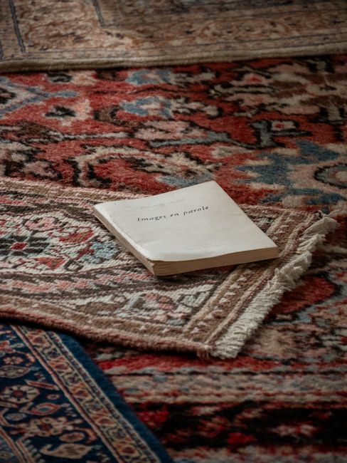 A person performing surface cleaning on a traditional patterned area rug in a living room, using a yellow vacuum cleaner with a black hose. The rug features intricate floral motifs in soft, muted colo